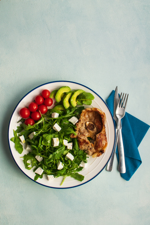 Diet menu. Healthy salad of fresh vegetables: tomatoes, avocado, arugula with cubes Goat cheese and Lamb chop in a plate. Space for text. Flat Lay. Top view. Vertical.の写真素材