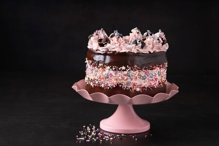 Birthday cake decorated with sprinkles chocolate ganache and pink color decor on a cake stand, dark background with copy space. Valentine's, Mother's Day. Trendy sprinkle fault line cake decor.の写真素材