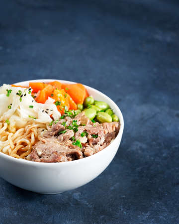 Poke bowl, pulled pork, vegetables, pumkin, soy beans edamame, noodles, on bowl, chopsticks. Copy space, closeup. Hawaiian dish, blue dark background. Trendy asian food. Healthy, clean eating concept.の写真素材