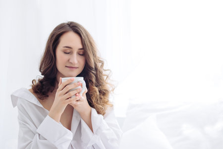 Portrait of young woman smelling coffee on white bed at morning. holding a cup. Breakfast, jeans, mans shirtの写真素材