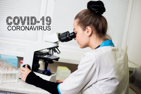 Covid-19 coronavirus poster. Young female doctor looking in a microscope in a clinic labalatory....
