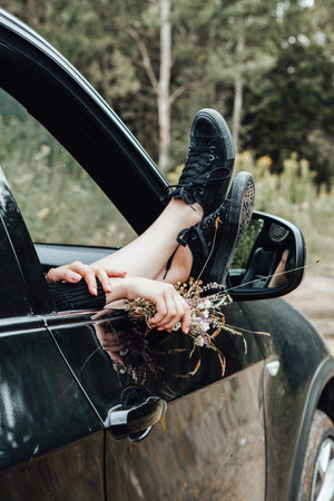 Woman feet in sneakers, open window, black car, forest, bouquet of wild flowers, early autumn.