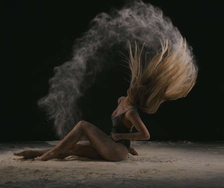 Beautiful sensual fit slim dancer in black body with long blond hair throwing dust, flour, powder on black background.の写真素材