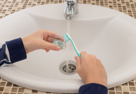 childrens hands take care of the removable orthodontic plate with toothbrush over the bathroom sink.