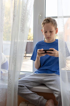 Preteen boy uses mobile phone while sitting on the windowsill at home.