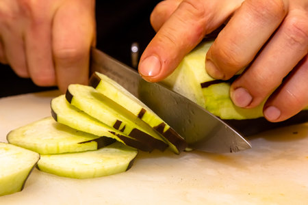 A professional chef quickly cuts eggplants on a wooden board using a ceramic knife. close-up.