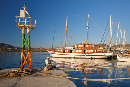 Sail boats and a man fishing on a pier next to harbour light in the port of Parikia, Paros island, Greeceのeditorial素材