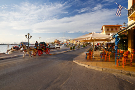View of the seafront with coffee shops, bars and restaurants and a carriage pulled by a horse at Aegina seafront, Greeceのeditorial素材