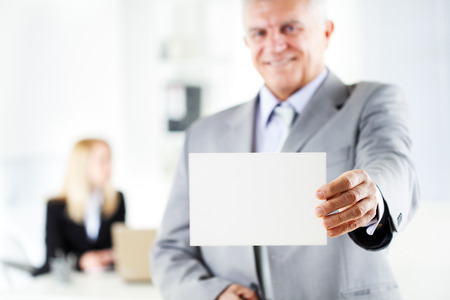 Happy senior businessman holding Blank business card in the office. Looking at camera. Selective Focus. Focus on business card.の写真素材