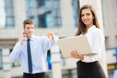 Young business couple standing in front of the building. Business woman carrying laptop while her partner use a mobile phone for business conversation.の写真素材