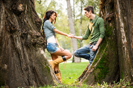 Young and pretty girl and guy at park leaning against a tree trunk, facing each other holding hands.の写真素材