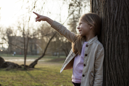 Smiling little girl in the park, leaning against a tree, showing something with an outstretched finger.の写真素材
