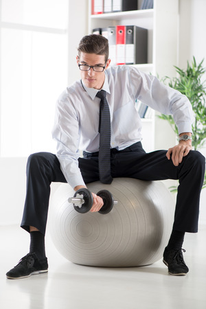 The young businessman sitting in the office on pilates ball and doing exercise with dumbbells.の写真素材