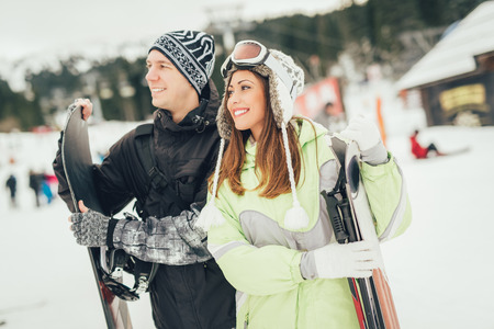Beautiful young couple enjoying in winter vacations. They standing with skis and snowboard and looking away.の写真素材