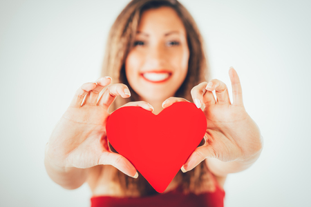 Close-up of a beautiful smiling girl holding a red heart. Selective focus. Focus on the heart.の写真素材