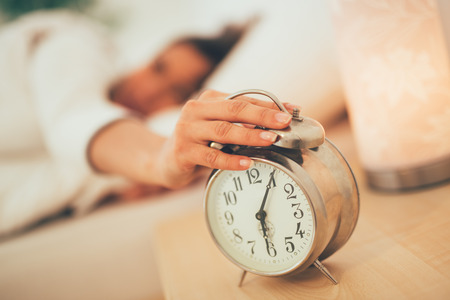 Beautiful young woman sleeping in bed and holding hand on alarm clock. Selective focus. Focus on foreground, on clock.の写真素材
