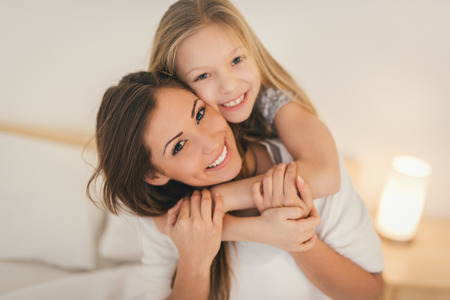Beautiful smiling mother and her daughter hugging in bed. They are sitting on bed at pajamas and smiling looking at camera.の写真素材