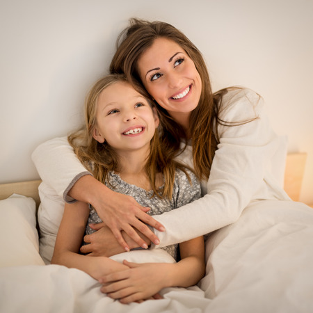 Beautiful smiling mother and her daughter hugging in bed. They are sitting on bed at pajamas and smiling.の写真素材