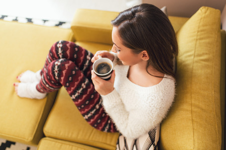 Beautiful young smiling woman relaxing on sofa at living room while enjoying in coffee time.の写真素材