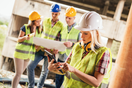 Beautiful young female construction architects using digital tablet at a construction site. Her colleagues review plan in background.の写真素材