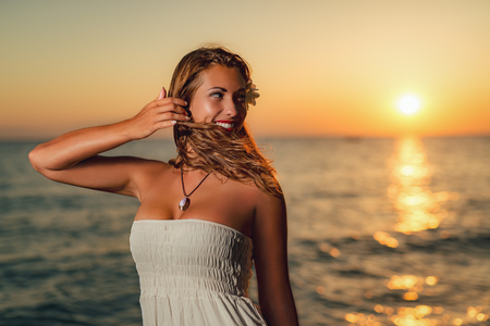 Portrait of a pretty young smiling woman in white dress enjoying sunset on the beach.の写真素材