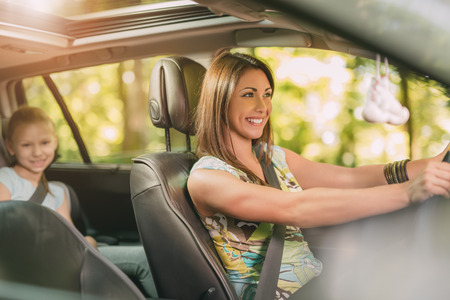 Young beautiful woman with a nice smile driving a car.  Her cute daughter sitting on rear and...