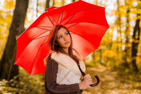 Portrait of a beautiful pensive young woman with red umbrella in sunny forest in autumn colors.の写真素材