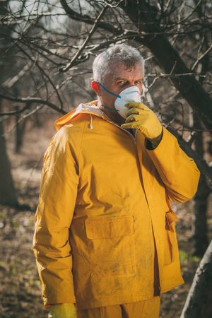 Mature man with mask spraying tree with chemicals in the orchard before winter. Looking at camera.の写真素材
