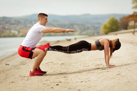 Young muscular couple having workout together on the beach. Handsome boy doing squats exercise, his cute girlfriend doing push-up.の写真素材