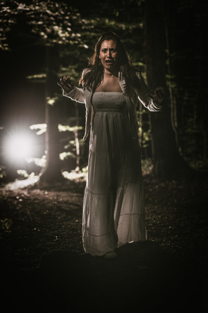 Young scared woman walking through the forest at night in white dress and screaming. Horror scene.の写真素材