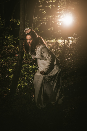 Young scared lonely woman walking through the forest at night in white dress.の写真素材