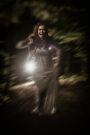 Young frightened woman running through the forest at night in white dress and screaming. Horror scene.の写真素材
