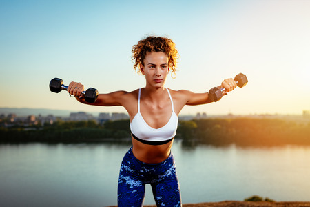 Young fitness woman doing workout with dumbbell by river on the city rooftop.
