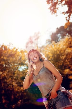 Beautiful smiling young woman with summer hat enjoying in the nature in autumn. She is walking and talking on smartphone.の写真素材