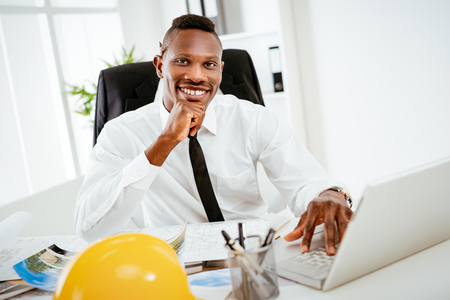 Smiling African construction engineer sitting at desk in the office and looking at camera. の写真素材