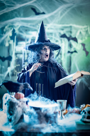 Witch with awfully face in creepy surroundings and smoky green background reading recipe of magic drink.の写真素材