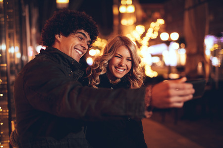 Young beautiful cheerful couple taking a selfie in the city street at new year's night with a lot of lights on background. の写真素材
