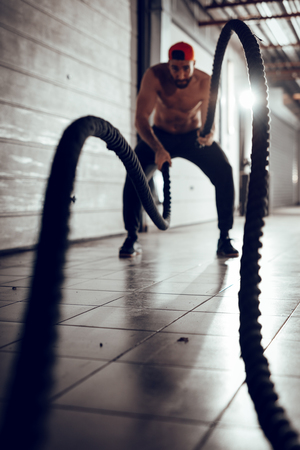 Handsome muscular young man exercising with ropes at the garage gym. Selective focus.の写真素材