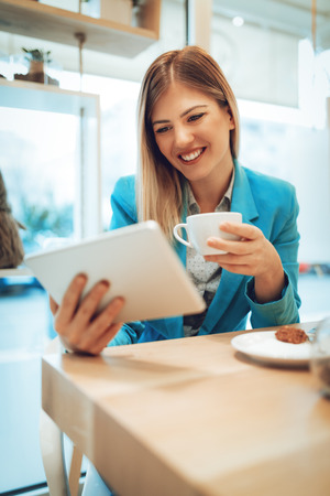 Beautiful young smiling woman checking social media on a digital tablet and drinking coffee in a cafe.の写真素材
