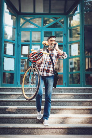 Casual businessman talking on smart phone and carrying the bike down the stairs to the entrance of the office building.の写真素材