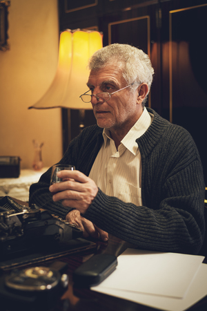 Retro senior man writer with glasses, sitting at the desk beside obsolete typewriter, with a glass of whiskey and thinking.の写真素材