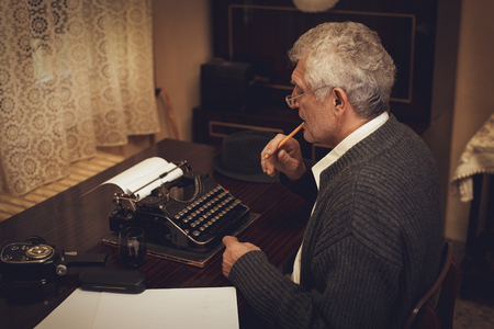 Retro senior man writer with glasses and pencil in his mouth sitting and looking on the text on obsolete typewriter.の写真素材