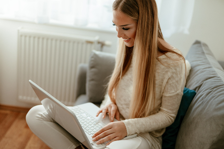 Beautiful young smiling woman relaxing at the home and surfing the net on a laptop.の写真素材