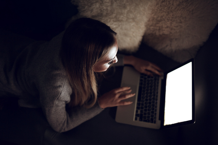 Cute young woman laying down on bed and using laptop. She is surfing on the net and checking social media.の写真素材