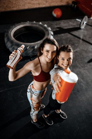 Two young smiling female friends resting after training at the gym. They are holding energy drinks and looking at camera. Selective focus. Top view.の写真素材