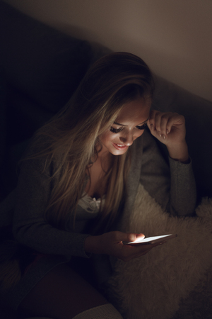 Cute girl sitting on bed and checking social media on smart phone at the night.の写真素材
