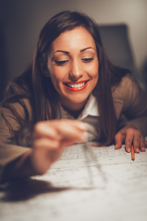Smiling young construction engineer analyzing blueprint at desk in office. She is measuring with drawing divider. Close-up. Selective focus. の写真素材
