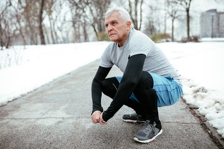 Active senior making pause crouching, during stretching and doing exercises in public park during the winter training outside in. Copy space.の写真素材
