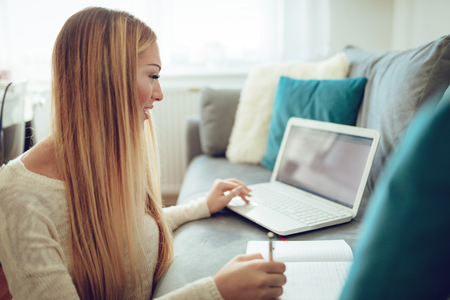 Beautiful young woman studying and working on her laptop and writing at the notebook, sitting at the floor beside the sofa at home.の写真素材
