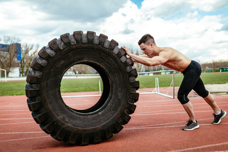 Young muscular man flipping a huge tire for training muscles at the tartan track on the stadium.の写真素材